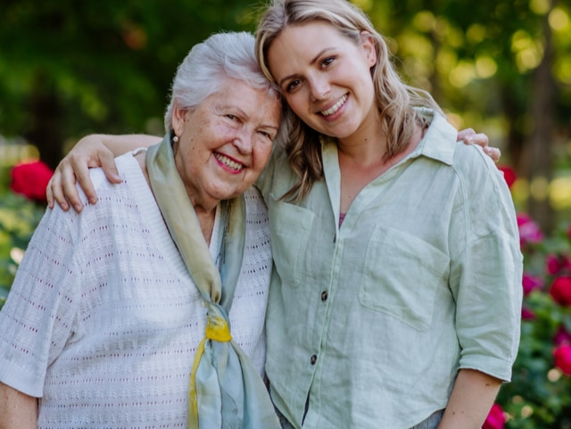 Smiling elderly woman and younger woman with arms around each other in a sunny garden with flowers in bloom.